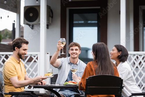 happy man raising glass of wine near interracial friends during bbq party on backyard