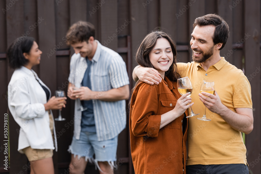 Fototapeta premium cheerful couple holding glasses with wine while hugging near blurred interracial friends on blurred background