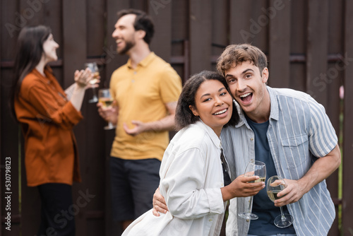 cheerful interracial couple holding glasses with wine near blurred friends on blurred background