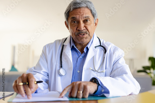 Senior biracial male doctor sitting at desk talking during video call consultation