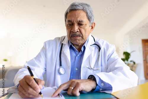 Senior biracial male doctor sitting at desk taking notes during consultation