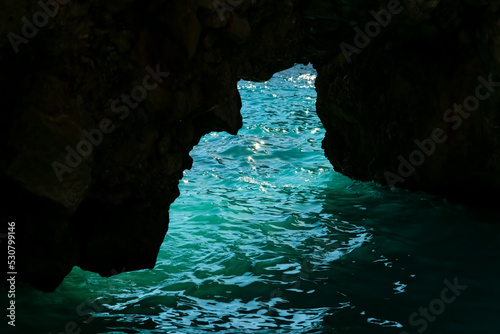 Small cave on the coast of Capri island Italy. Turquoise green to blue water reflecting sunlight and blue sky. Silhouette of rock formations on the shore of “Marina piccola“ a popular beach site.