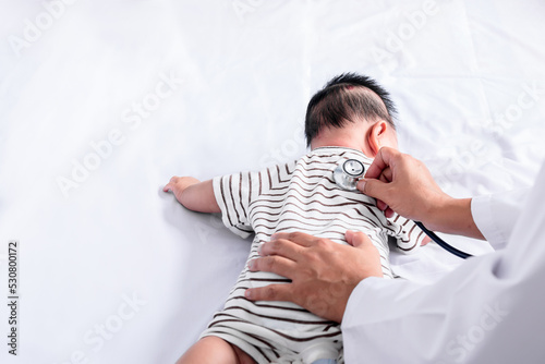 Doctor using a stethoscope checking the respiratory system and heartbeat of a 3-months-old an Asian baby newborn, is lying on white bed, to health care and baby newborn concept.
