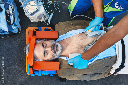 Ambulance paramedic using defibrillator performing cardiopulmonary resuscitation on casualty. First aid for injured mature man lying on emergency stretcher, top view