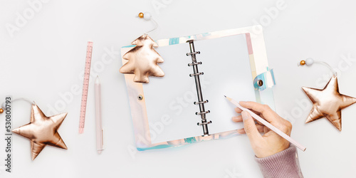 Female writing in copybook or diary with bright cover on white background, shiny Christmas tree and star. Concept of planning personal future goals and ideas for New Year, top view banner