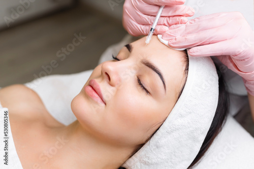Close up of hands of young cosmetologist injecting botox in female face. She is standing and smiling. The woman is closed her eyes with relaxation photo