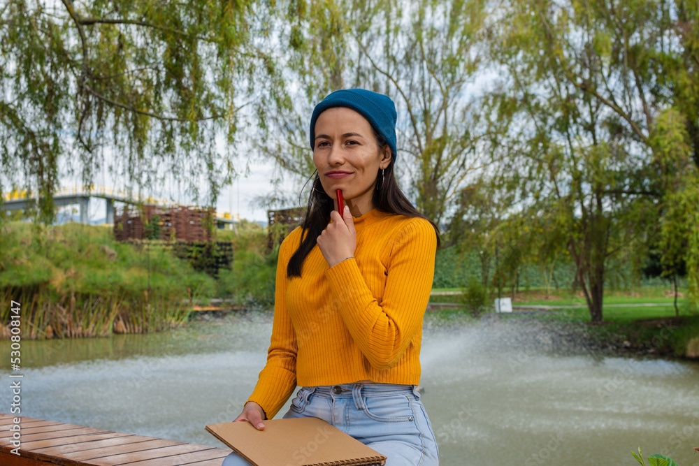 Latina student woman wearing yellow sweater and blue hat outdoors with a thinking expression while holding a notebook and a pen Latina student woman wearing yellow sweater and blue hat outdoors with a thinking expression while holding a notebook and a pen