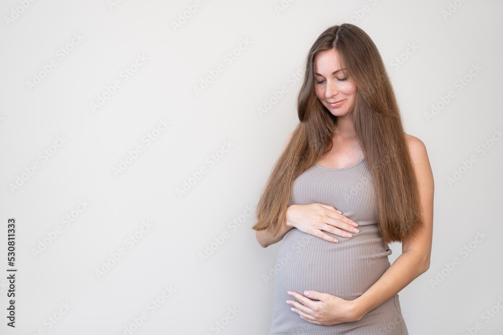 Happy attractive pregnant woman, expecting child wearing tight dress looking at her belly on gray background. Pregnancy, love, motherhood concept.Copy space. Happy attractive pregnant woman, expecting child wearing tight dress looking at her belly on gray background. Pregnancy, love, motherhood concept.Copy space.