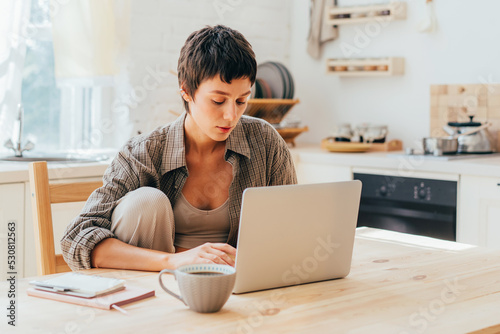 Young short-haired modern woman working  on the computer while sitting at the table in the kitchen