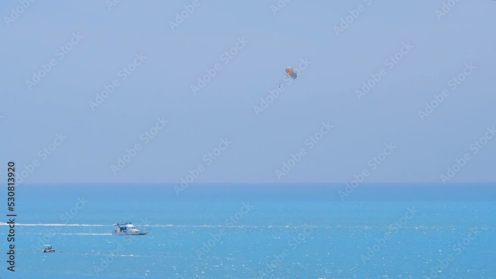 Multicolor parachute is hiding behind the boat. Parasailing, active form of recreation. A sailboat, a speedboat, a jet ski, a catamaran sailing on the blue sea leave white traces behind them. Multicolor parachute is hiding behind the boat. Parasailing, active form of recreation. A sailboat, a speedboat, a jet ski, a catamaran sailing on the blue sea leave white traces behind them.