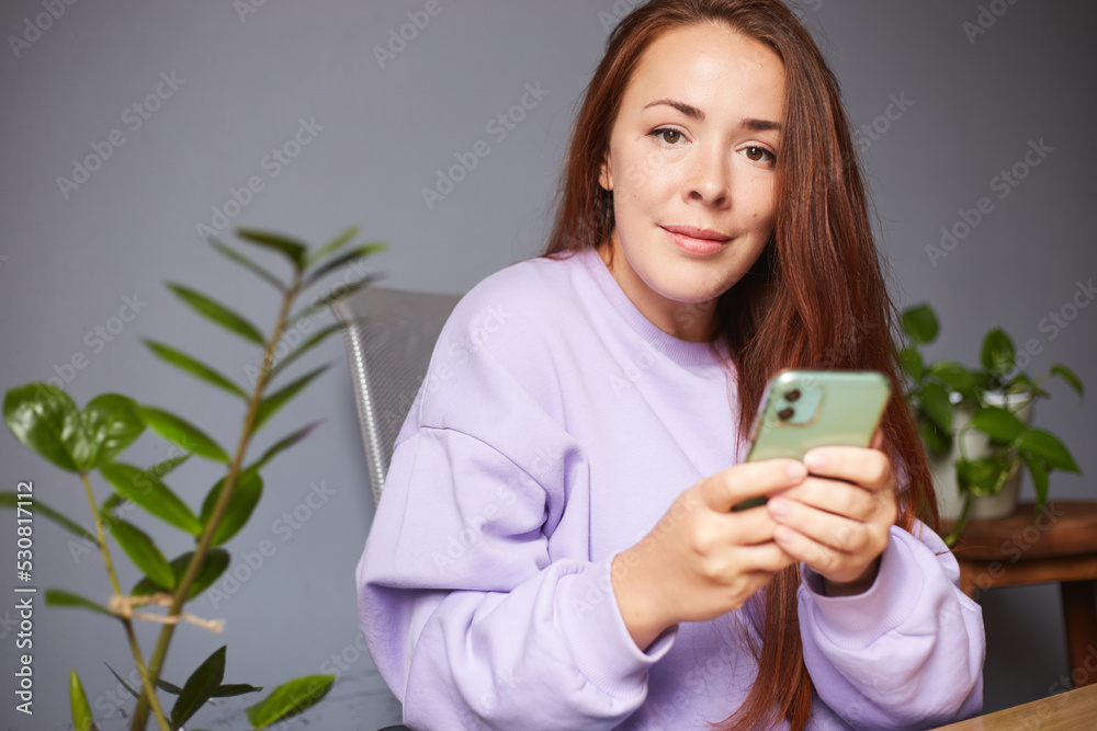 Cheerful caucasian female use her smartphone. A lot of green plants around. Red hair woman in violet sweater Cheerful caucasian female use her smartphone. A lot of green plants around. Red hair woman in violet sweater