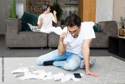 Young husband sitting on the floor with many bills and checks and calculating expenses of his spender wife who sitting on the couch and relaxing photo