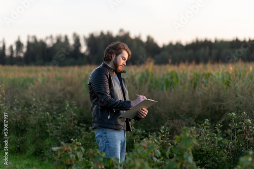 Young farmer writes notes while standing against the background of a blurred corn field © Zelma