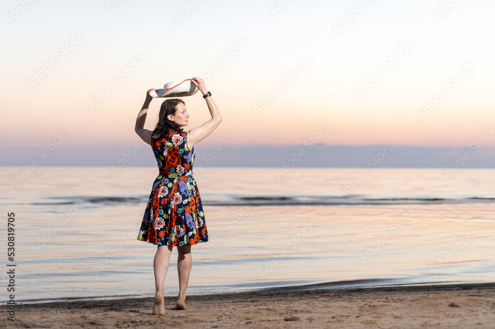 Happy young woman in flowery dress and summer hat dancing joyfully along seashore Happy young woman in flowery dress and summer hat dancing joyfully along seashore