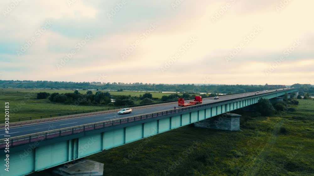 Red powerful truck with trailer for loading heavy construction technics driving over bridge at sunset. Concept of optimizing cargo transportation business Red powerful truck with trailer for loading heavy construction technics driving over bridge at sunset. Concept of optimizing cargo transportation business
