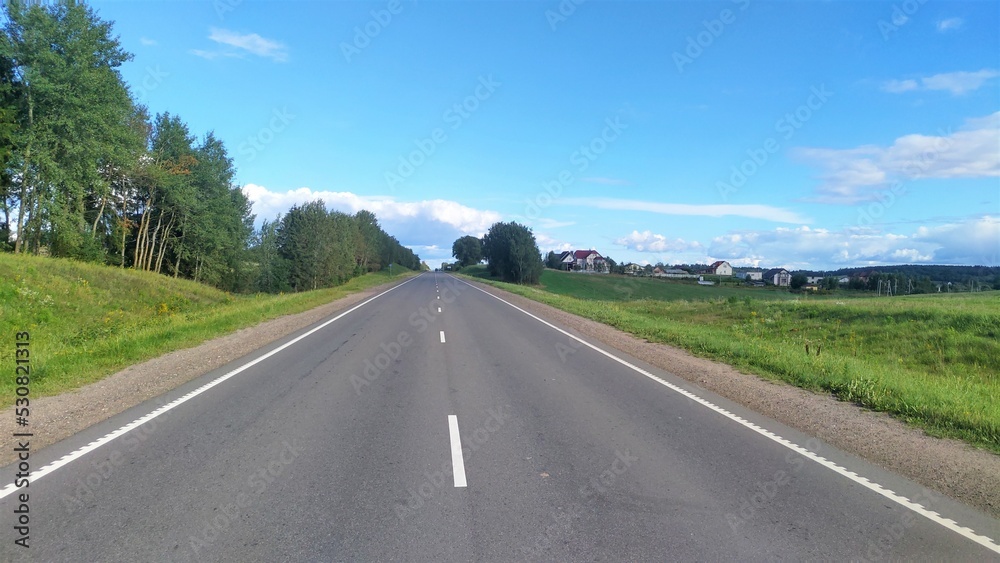 An asphalt road with markings and sandy shoulders passes among grassy meadows. The road is flanked by trees. In the distance is a village. Sunny weather and blue skies with clouds