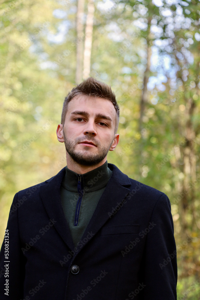 Handsome guy in a black coat in the forest. Cute guy with stubble and a model hairstyle is posing in the forest while standing in a coat. Handsome guy in a black coat in the forest. Cute guy with stubble and a model hairstyle is posing in the forest while standing in a coat.