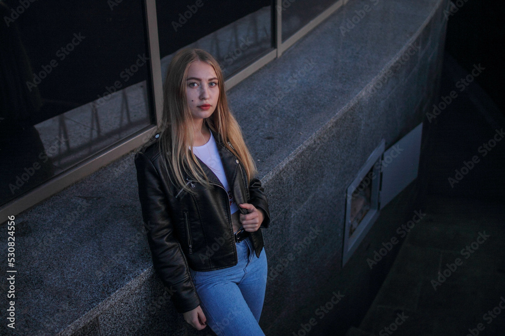 a blonde girl in a leather jacket and light jeans stands near modern building and looking into camera a blonde girl in a leather jacket and light jeans stands near modern building and looking into camera