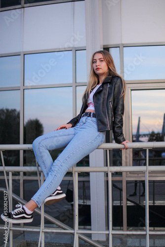 a young beautiful blonde girl is sitting on the railing near a new modern building in a leather jacket and jeans in autumn