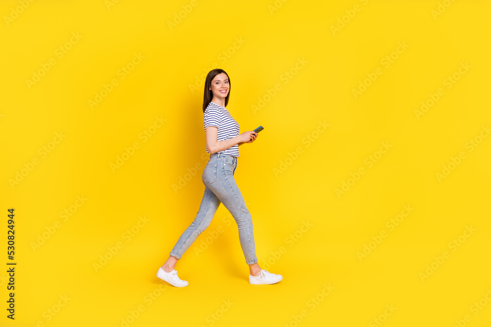 Full size profile photo of satisfied lady walking empty space arm hold use modern device gadget isolated on yellow color background