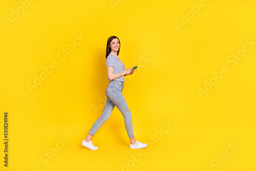 Full size profile photo of satisfied lady walking empty space arm hold use modern device gadget isolated on yellow color background