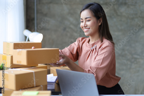 Young Asian woman start up business owner sales, working on laptop and cardboard boxes to prepare orders for delivery.