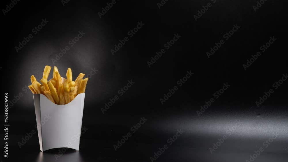 White paper packaging made of cardboard rotates around itself. French fries, fried, in a container. Fast food, production concept. Black background. A place to insert text. Copy space. 