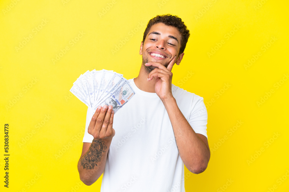Young brazilian man taking a lot of money isolated on yellow background thinking an idea while looking up Young brazilian man taking a lot of money isolated on yellow background thinking an idea while looking up