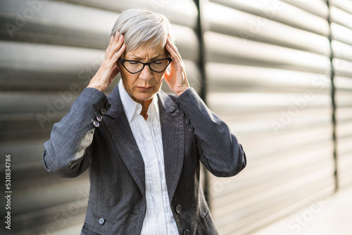 Senior businesswoman is having headache. She is standing in front of company building.