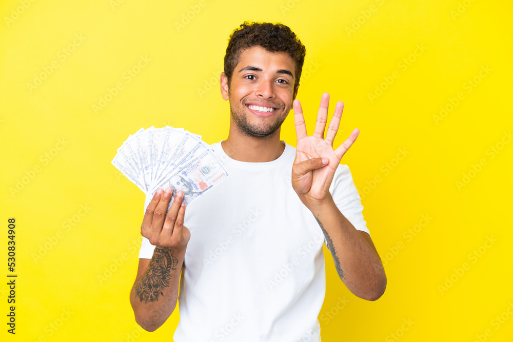 Young brazilian man taking a lot of money isolated on yellow background happy and counting four with fingers Young brazilian man taking a lot of money isolated on yellow background happy and counting four with fingers