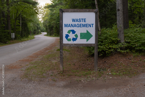 Sign in English waste management with a recycling symbol and a direction arrow by the side of rural road going through the forest. Selective focus, blurred background.