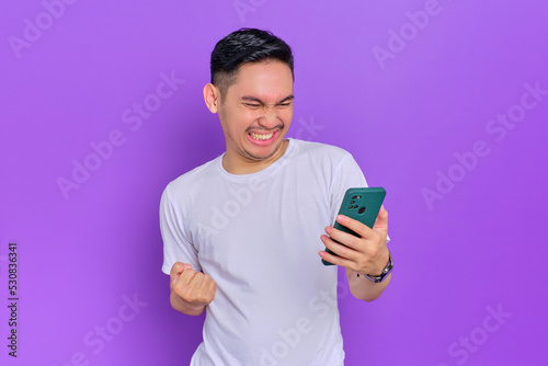 Excited young Asian man in white t-shirt using smartphone and making winner gesture isolated on purple background © Sewupari Studio
