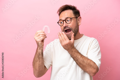 Middle age man holding invisible braces isolated on pink background with surprise and shocked facial expression