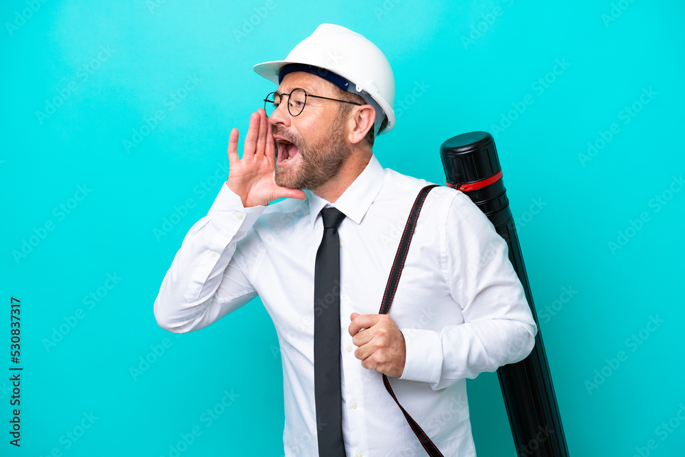 Middle age architect man with helmet and holding blueprints isolated on blue background shouting with mouth wide open to the side Middle age architect man with helmet and holding blueprints isolated on blue background shouting with mouth wide open to the side