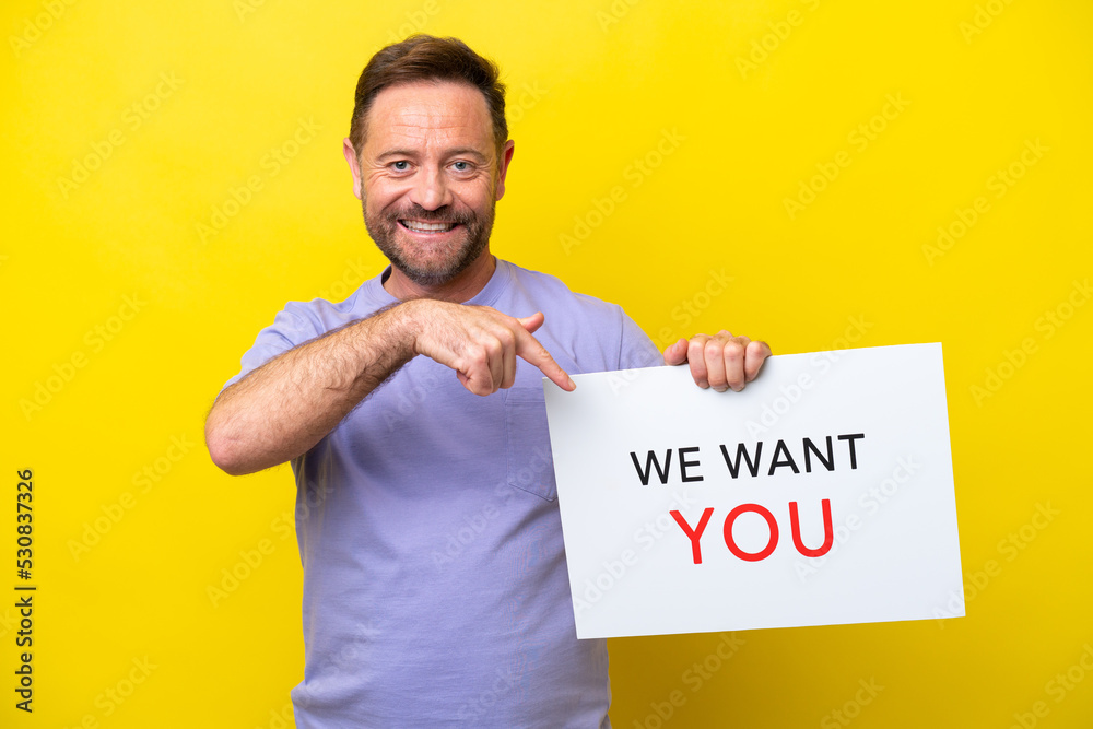 Middle age caucasian man isolated on yellow background holding We Want You board and  pointing it