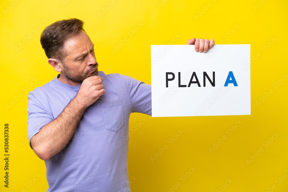 Middle age caucasian man isolated on yellow background holding a placard with the message PLAN A and thinking Middle age caucasian man isolated on yellow background holding a placard with the message PLAN A and thinking