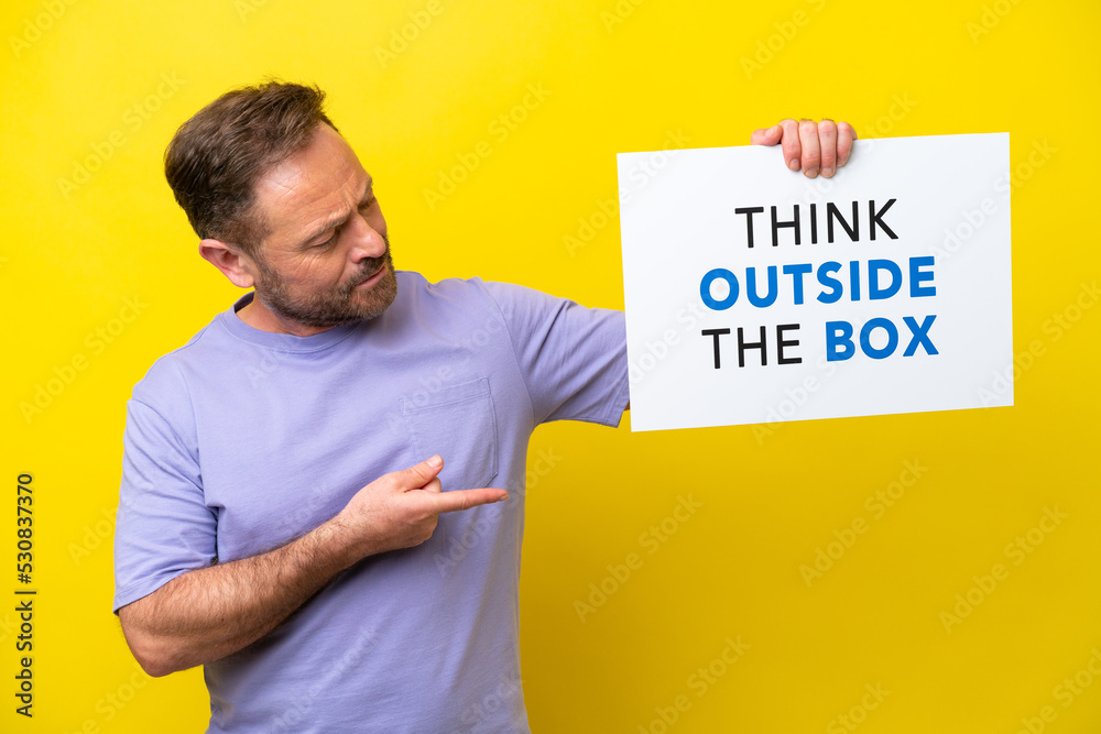 Middle age caucasian man isolated on yellow background holding a placard with text Think Outside The Box and pointing it Middle age caucasian man isolated on yellow background holding a placard with text Think Outside The Box and pointing it