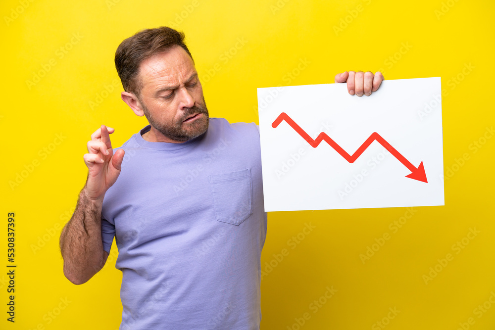 Middle age caucasian man isolated on yellow background holding a sign with a decreasing statistics arrow symbol with fingers crossing Middle age caucasian man isolated on yellow background holding a sign with a decreasing statistics arrow symbol with fingers crossing