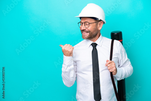 Middle age architect man with helmet and holding blueprints isolated on blue background pointing to the side to present a product