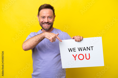 Middle age caucasian man isolated on yellow background holding We Want You board and  pointing it © luismolinero