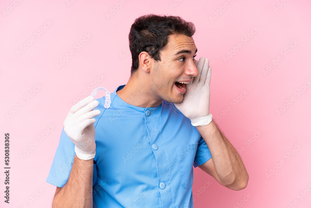 Dentist caucasian man holding invisible braces isolated on pink background shouting with mouth wide open to the side Dentist caucasian man holding invisible braces isolated on pink background shouting with mouth wide open to the side