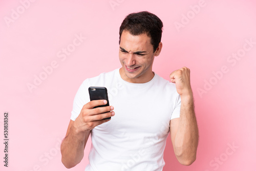 Young caucasian man isolated on pink background using mobile phone and doing victory gesture © luismolinero