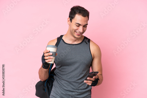 Young sport caucasian man with sport bag isolated on pink background holding coffee to take away and a mobile