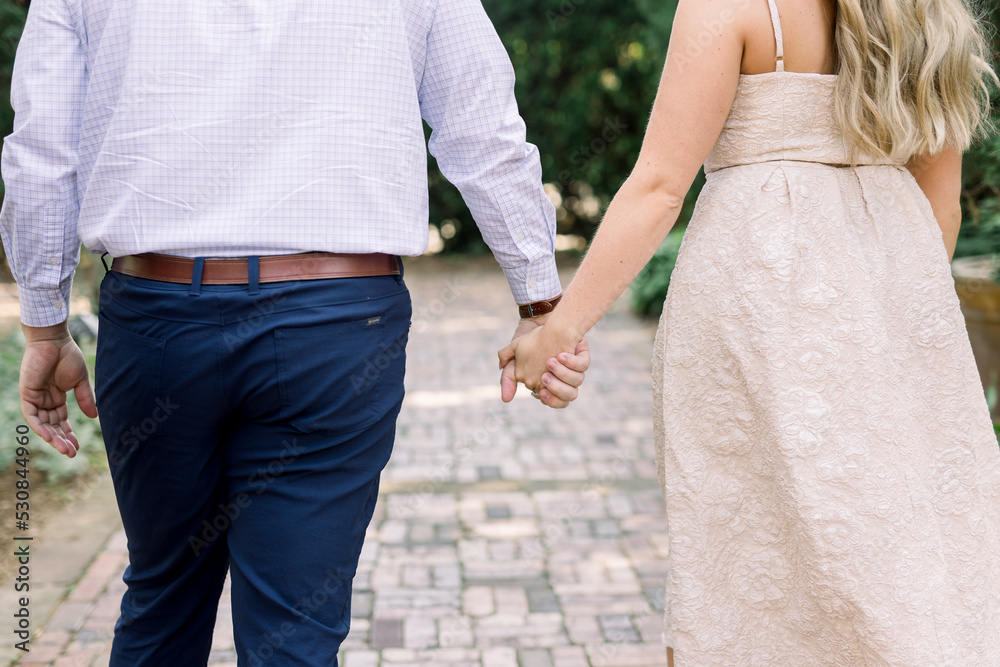 An engaged couple are holding hands and walking away. She is wearing a cream dress, and he has a dress shirt, belt, and pants on An engaged couple are holding hands and walking away. She is wearing a cream dress, and he has a dress shirt, belt, and pants on