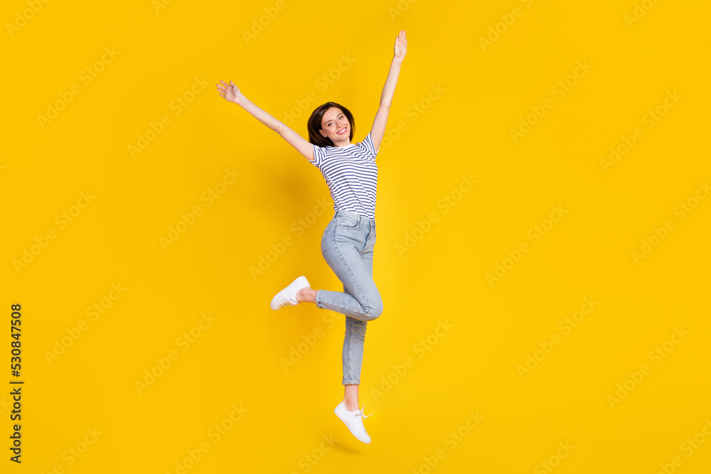 Full length photo of shiny young woman raise hands jumping high good mood dressed trendy striped look isolated on yellow color background