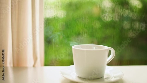 Wallpaper Mural Hot coffee or tea on a saucer. White ceramic hot coffee cup with smoke on the table in the living room by the window with white curtains. Outside, it was raining in the midst of a fresh green garden. Torontodigital.ca