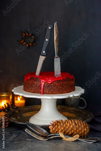 Moist chocolate cake with red bloody icing and two knives. Spider in the background, halloween mood and candles.