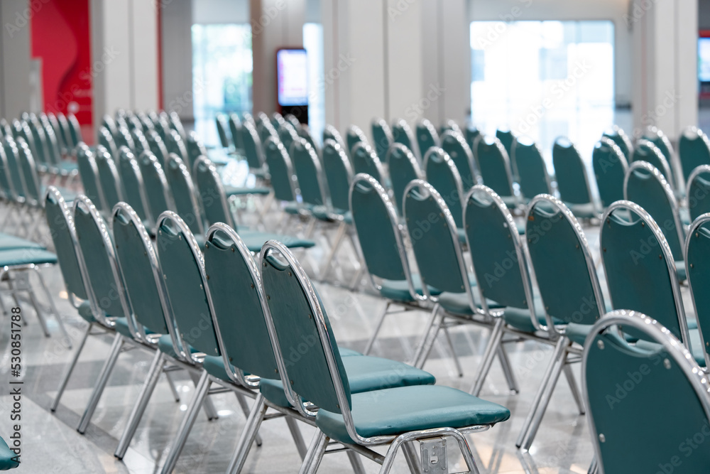 Many empty green leather chairs in a wide hall room, lights on. A floor reflect the objects. Give a feeling of emptiness.