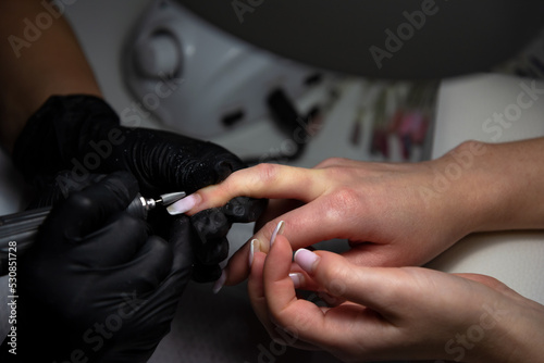 Close-up of a professional beautician removes old varnish with an electric drill. Hands of a manicure master with a tool.