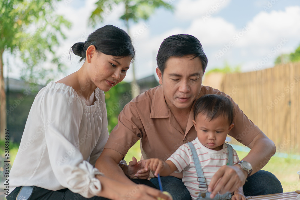 Fototapeta premium Asian family with baby little son painting eggs with brush in class workshop. Happy Asian father and mother with little son and teaching painting eggs with water colour